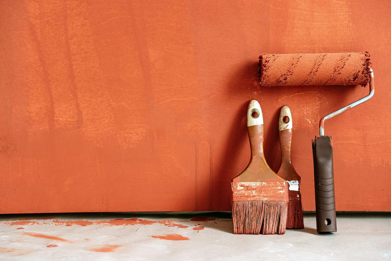 Close-up of paint rollers and brushes with orange paint against a painted wall.